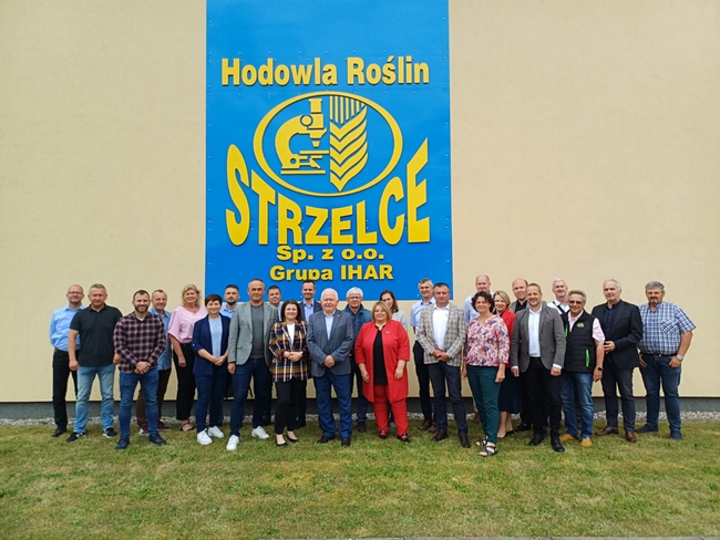 Wspólna fotografia uczestników Posiedzenia PDO. Stoją na zewnątrz, na trawie, przed budynkiem. Za nimi napis „Hodowla Roślin Strzelce sp. z o.o. Grupa IHAR